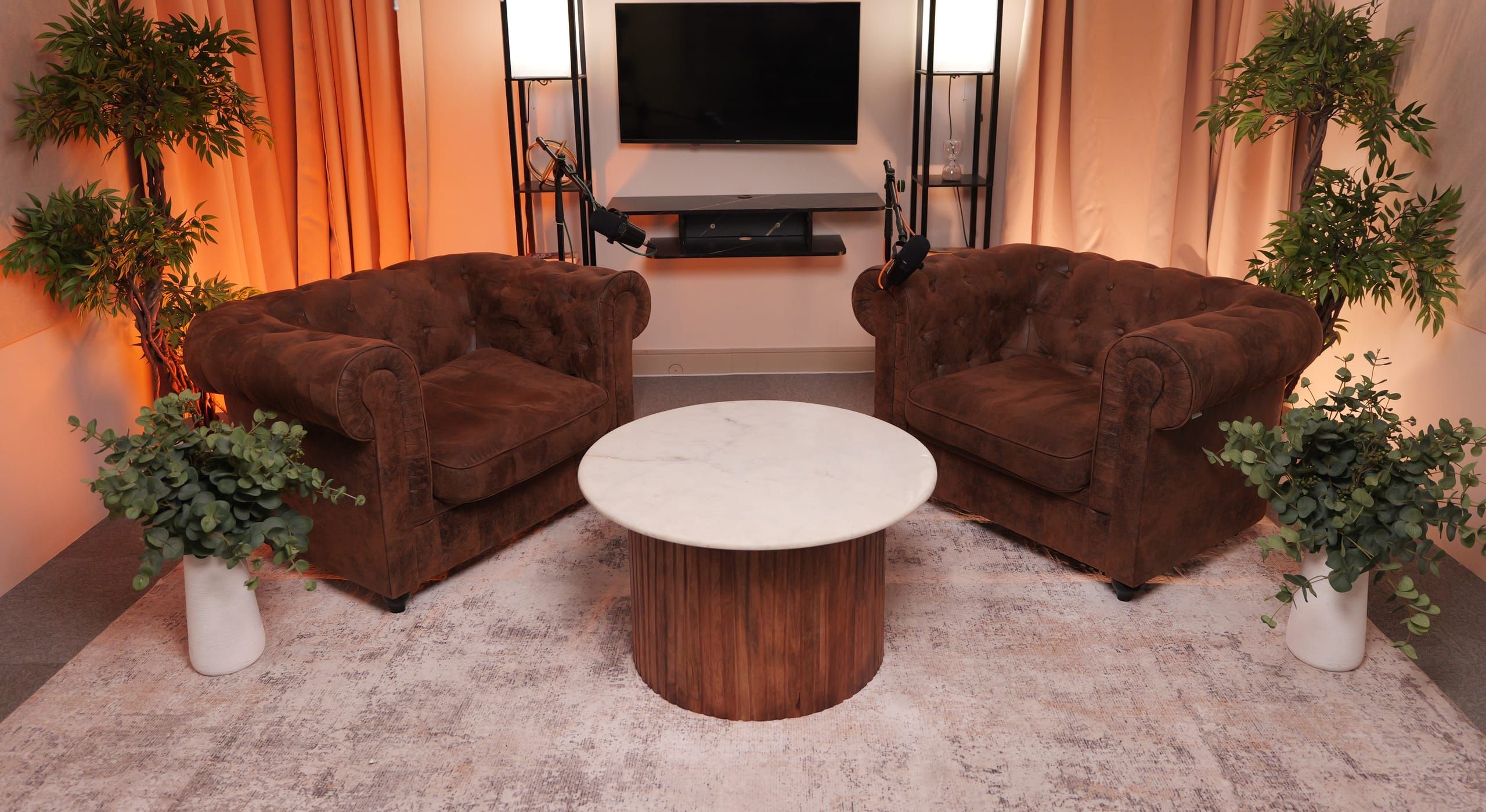 Room 1 at Myriad Studios: two Chesterfield armchairs angled around a marble and walnut table, between potted trees and warm lit curtains, with a microphone ready at each seat